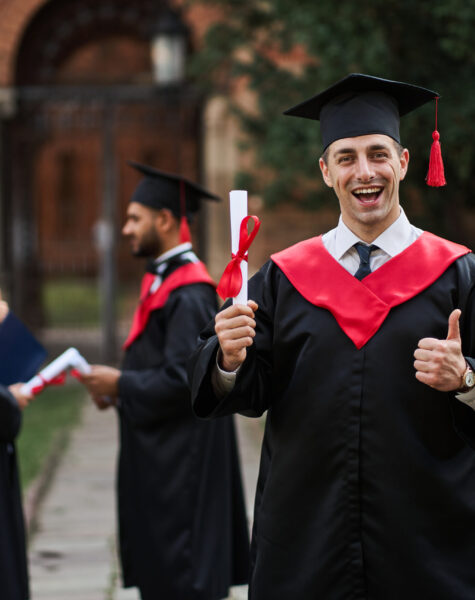 Happy caucasian graduate with his classmates in graduation gown holds diploma in campus.