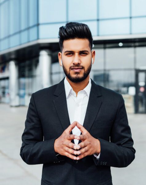 Portrait of a serious smiling modern Indian man, businessman, in front of the holding, behind him a modern building, he is looking at the camera.