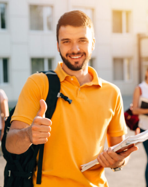 Young attractive smiling student showing thumb up outdoors on campus at the university. Selective focus